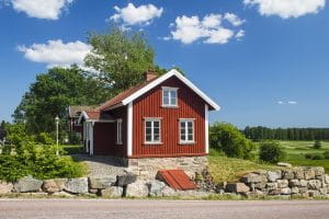 Et svensk, rødt træhus i med grønne træer og blå himmel som baggrund.