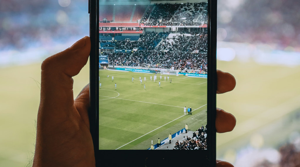 Iemand die in een stadion een voetbalwedstrijd volgt via zijn smartphone