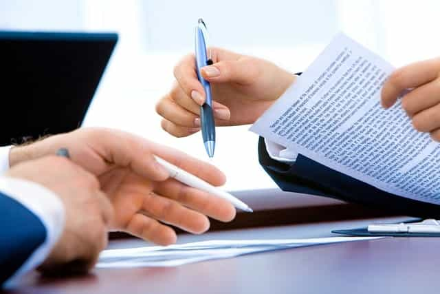 Two different individuals’ hands holding pens and reviewing a written contract before signing it.