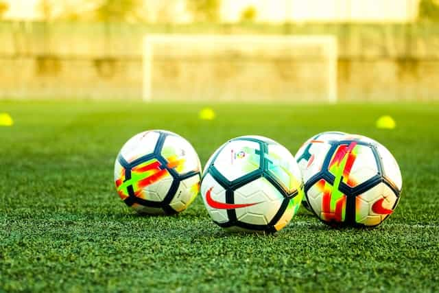 Three soccer balls sit on a green field with a net in the distance behind them.