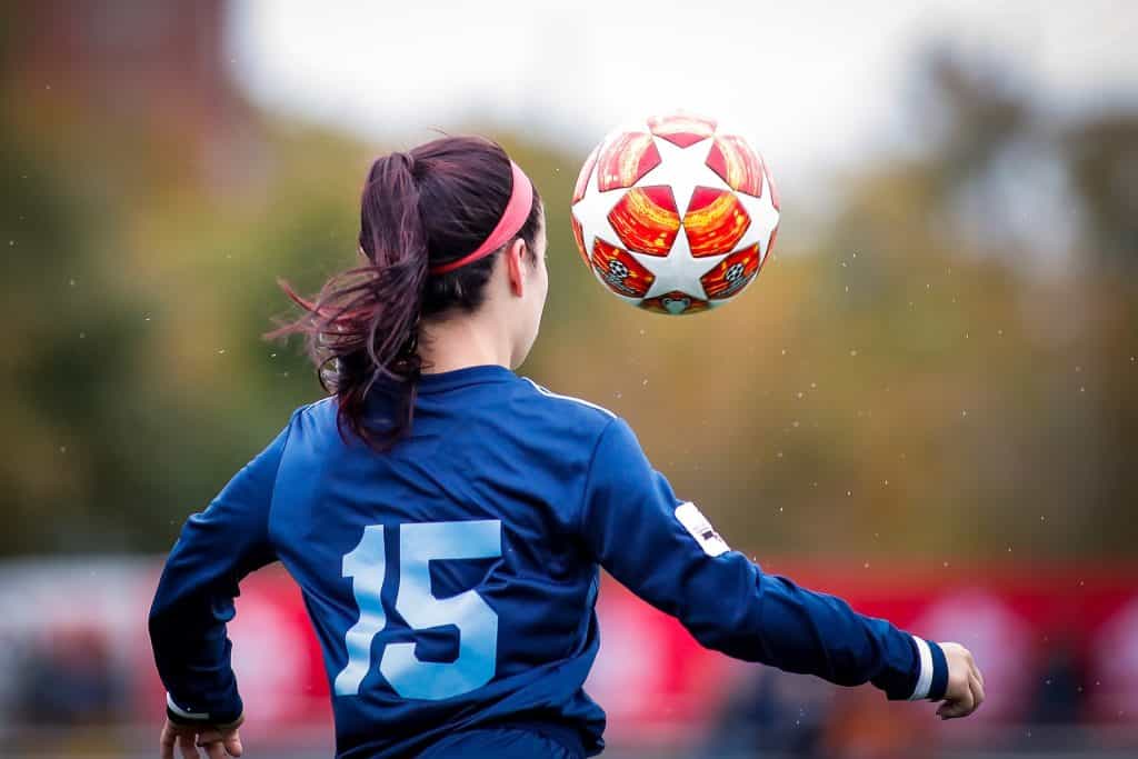 From behind, a woman kicks a red soccer ball high in the air while her ponytail swings.