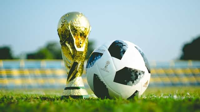 The golden World Cup trophy sits on a soccer field next to a soccer ball.